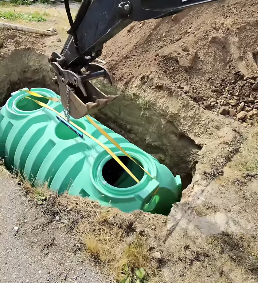 Excavator lowering a green septic tank into a prepared excavation.